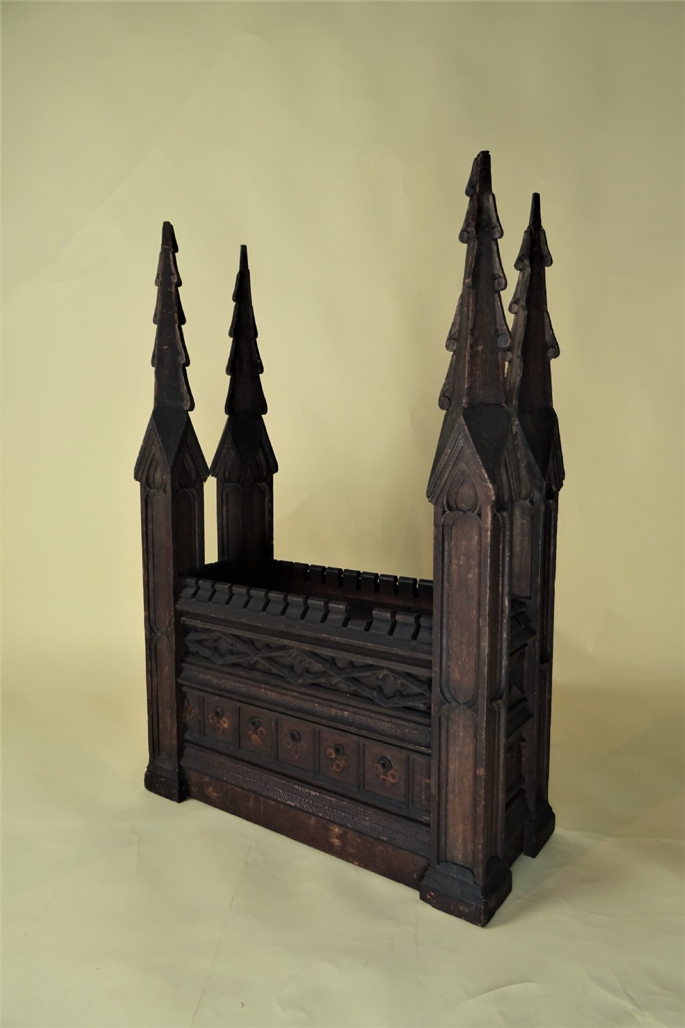 A dark carved oak Gothic architectural element featuring four corner spires with crocketed details, crenellated upper edges, and geometric tracery relief panels. View XIII