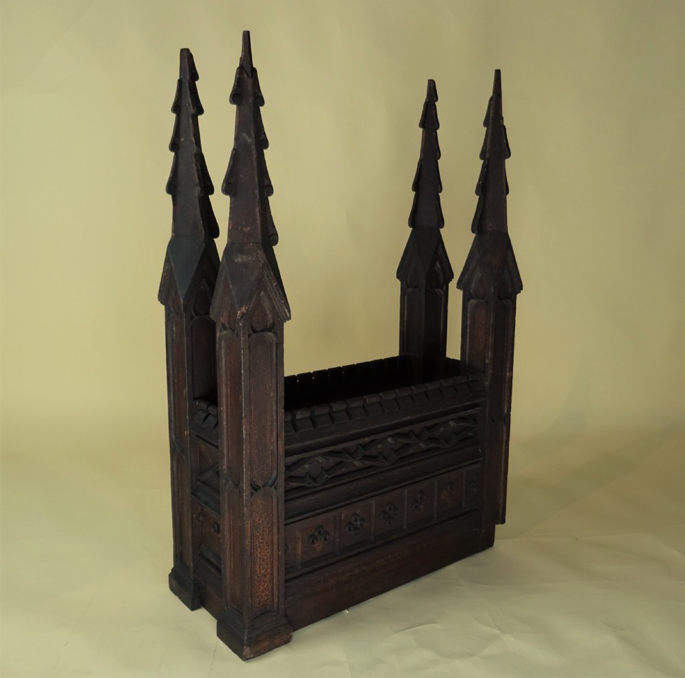 A dark carved oak Gothic architectural element featuring four corner spires with crocketed details, crenellated upper edges, and geometric tracery relief panels. View III