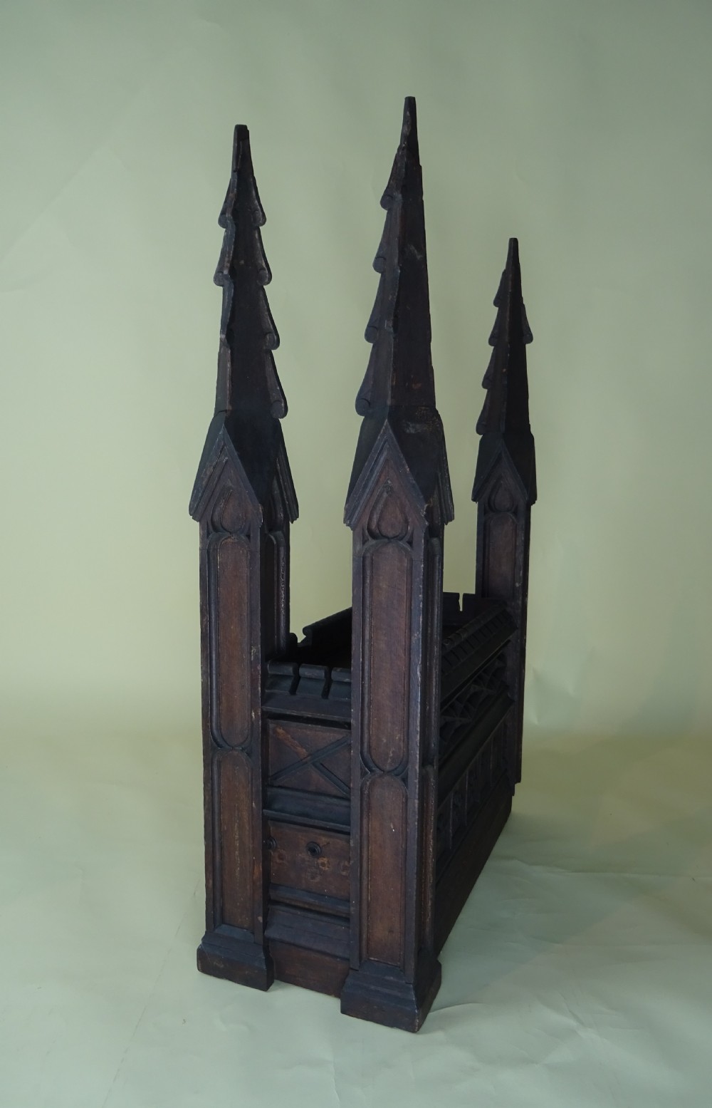 A dark carved oak Gothic architectural element featuring four corner spires with crocketed details, crenellated upper edges, and geometric tracery relief panels. View V