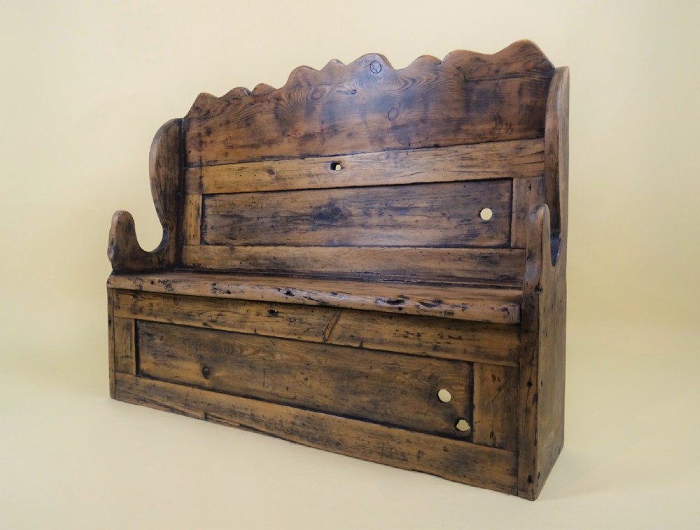 A miniature antique wooden pine settle featuring a scalloped top crest, solid seat, shaped winged sides, and a rustic weathered patina. View X