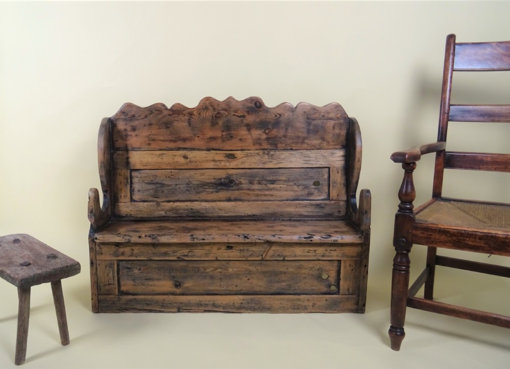 A miniature antique wooden pine settle featuring a scalloped top crest, solid seat, shaped winged sides, and a rustic weathered patina. View VI