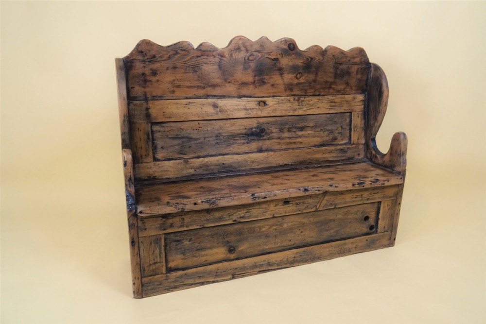 A miniature antique wooden pine settle featuring a scalloped top crest, solid seat, shaped winged sides, and a rustic weathered patina. View II