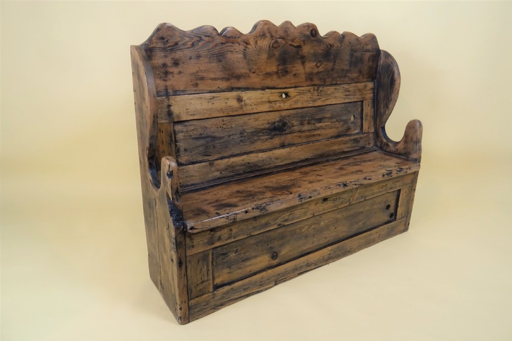 A miniature antique wooden pine settle featuring a scalloped top crest, solid seat, shaped winged sides, and a rustic weathered patina.