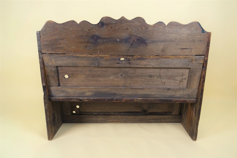 A miniature antique wooden pine settle featuring a scalloped top crest, solid seat, shaped winged sides, and a rustic weathered patina. View VIII