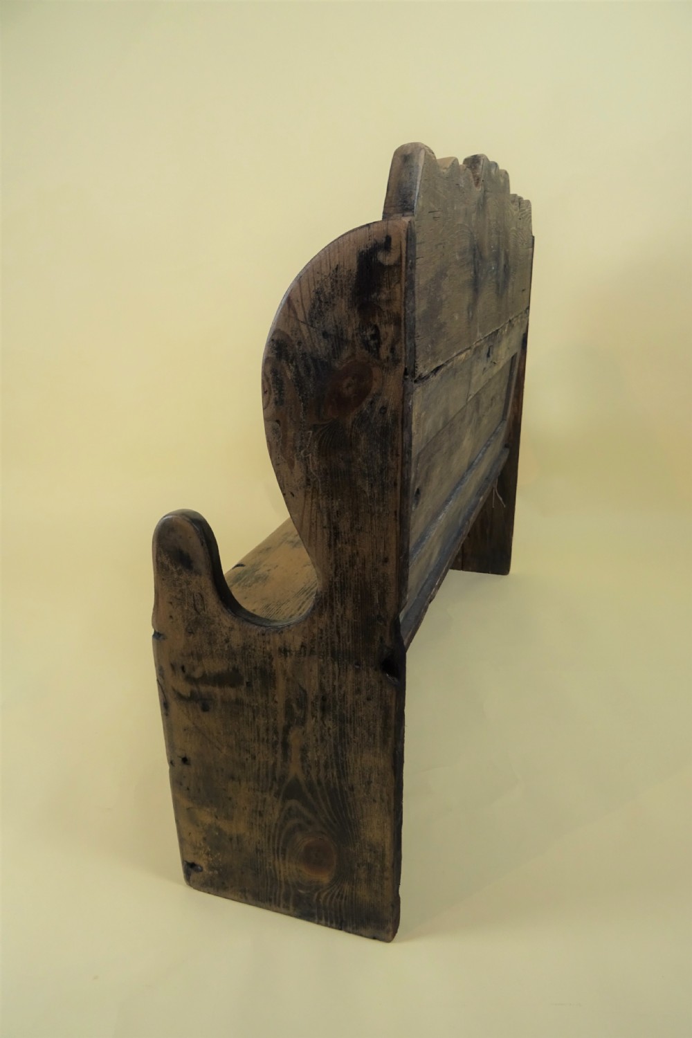 A miniature antique wooden pine settle featuring a scalloped top crest, solid seat, shaped winged sides, and a rustic weathered patina. View V