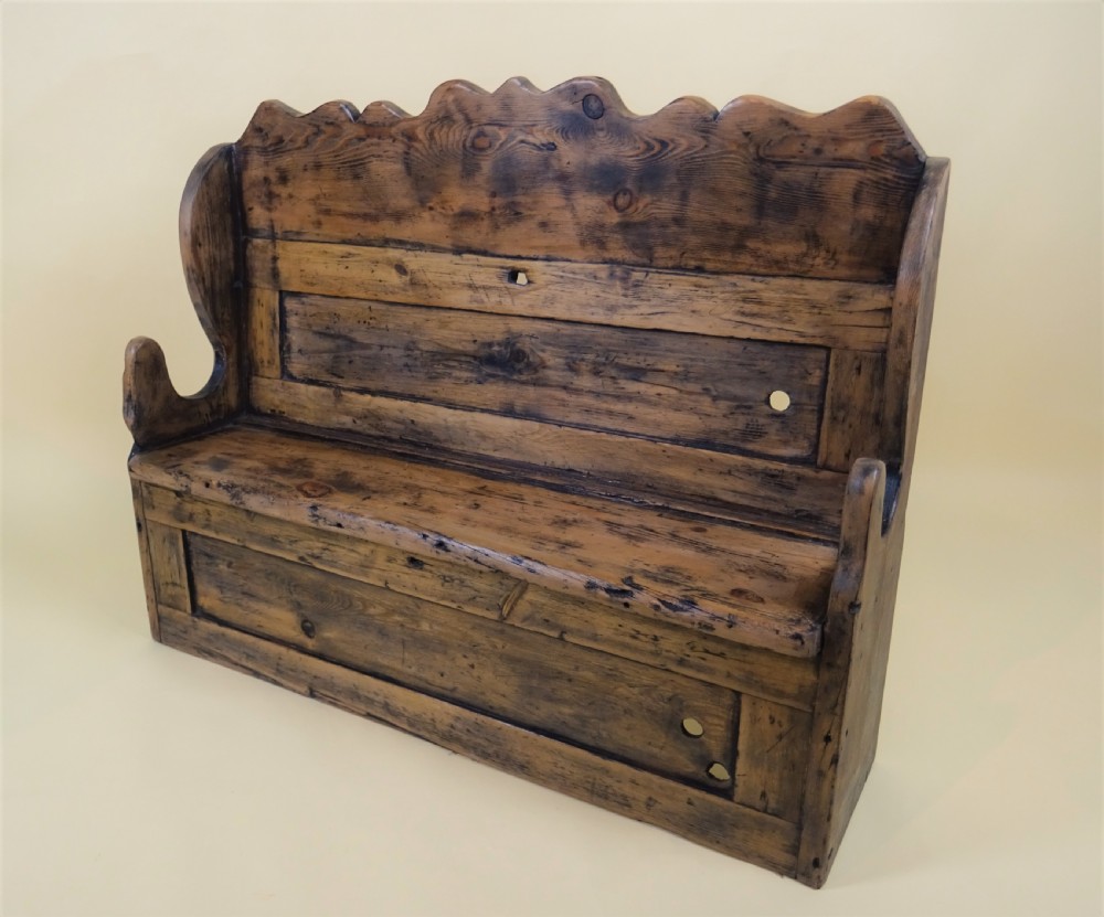 A miniature antique wooden pine settle featuring a scalloped top crest, solid seat, shaped winged sides, and a rustic weathered patina. View IX