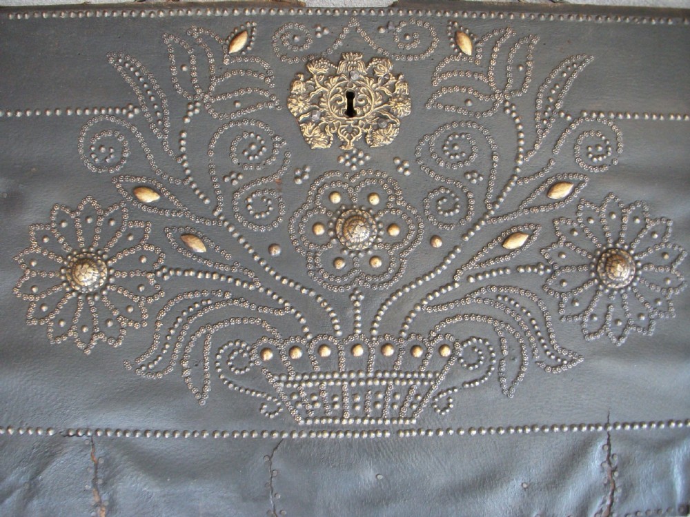 A rectangular dark leather-clad wooden panel adorned with floral and scroll patterns rendered in brass nailhead studs around a central brass escutcheon. View V