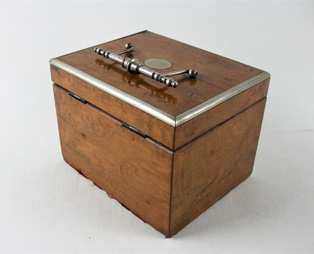 A rectangular antique burr maple wooden tea caddy featuring metal trim, a lid handle, an oval central cartouche, and a keyhole escutcheon. View V