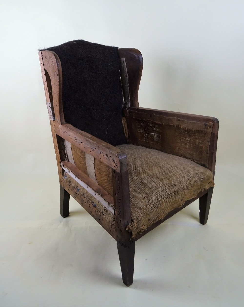 An antique childs wingback chair displaying an exposed wooden frame, original hessian under-upholstery, and dark back padding. View XI