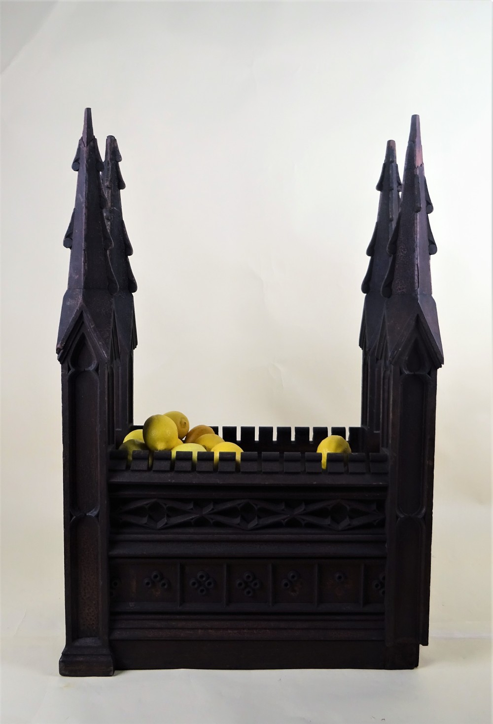 A dark carved oak Gothic architectural element featuring four corner spires with crocketed details, crenellated upper edges, and geometric tracery relief panels.