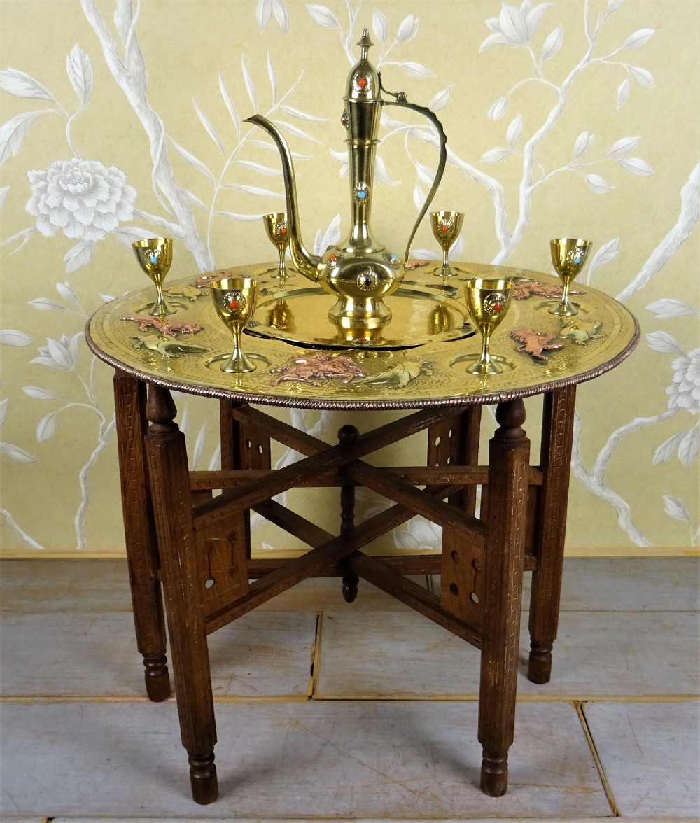 A circular engraved brass tray table with copper reliefs set upon a folding wooden base with a matching brass ewer and goblets.