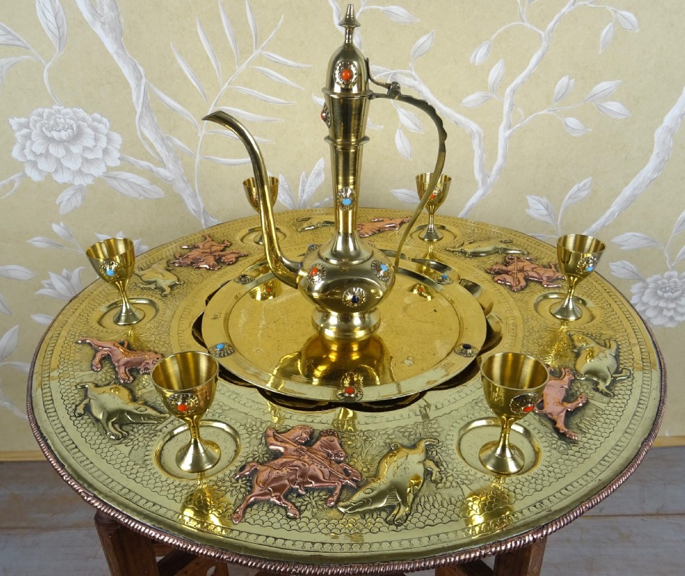 A circular engraved brass tray table with copper reliefs set upon a folding wooden base with a matching brass ewer and goblets. View X
