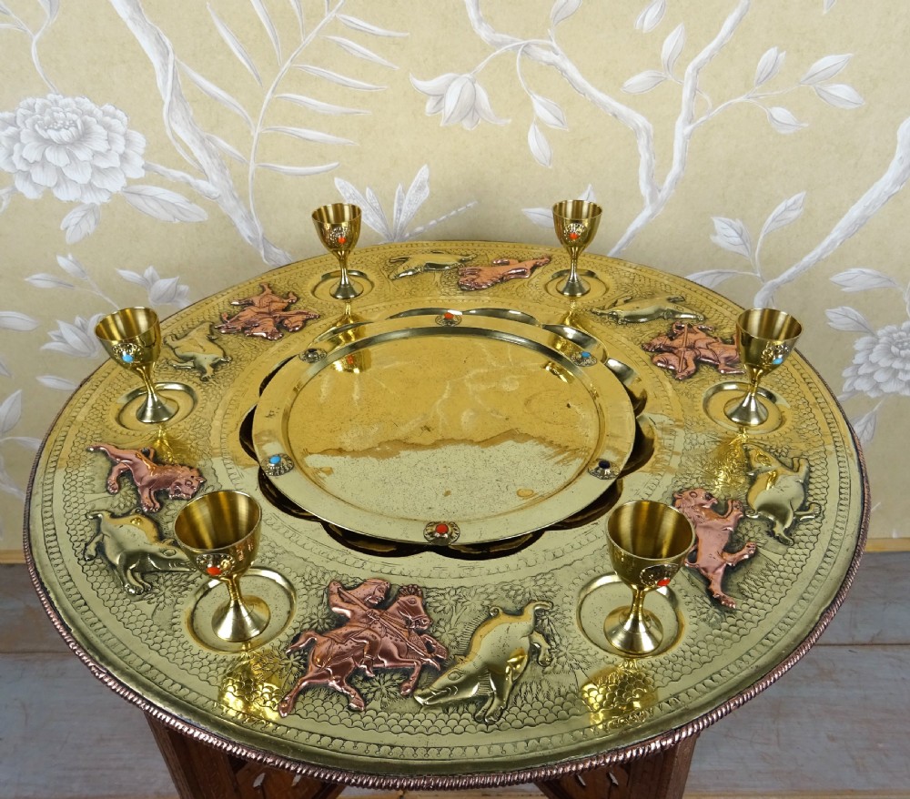 A circular engraved brass tray table with copper reliefs set upon a folding wooden base with a matching brass ewer and goblets. View II