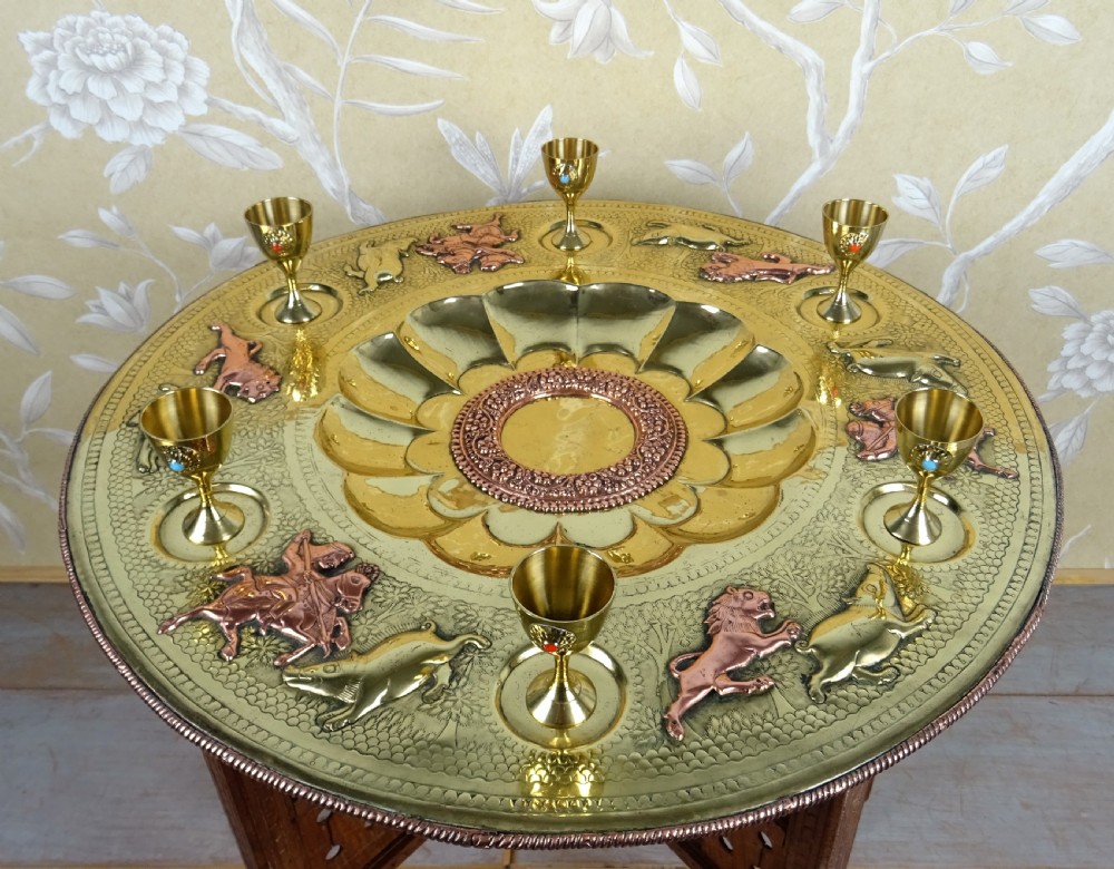 A circular engraved brass tray table with copper reliefs set upon a folding wooden base with a matching brass ewer and goblets. View VII