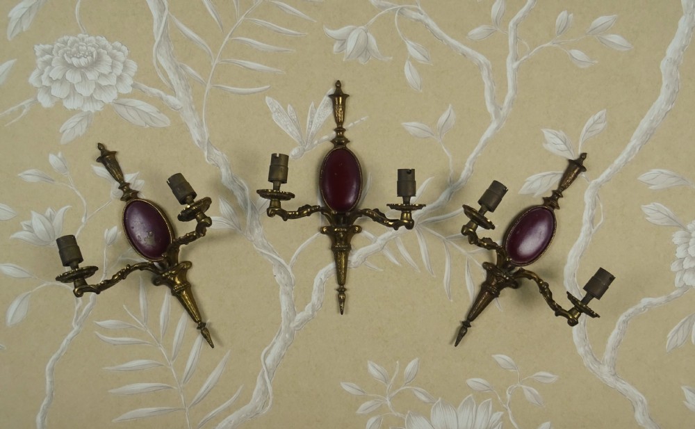 A set of three Regency style twin-arm brass wall sconces featuring oval red cabochons and decorative urn finials. View VII