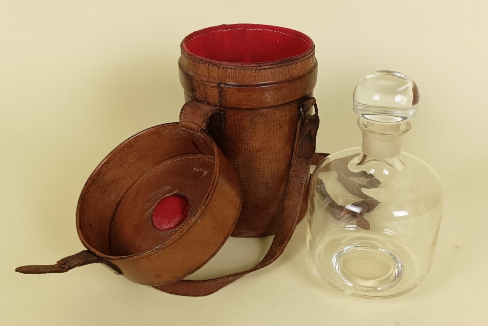 A clear glass decanter with a round glass stopper standing beside a cylindrical brown leather travelling case with a red fabric lining.