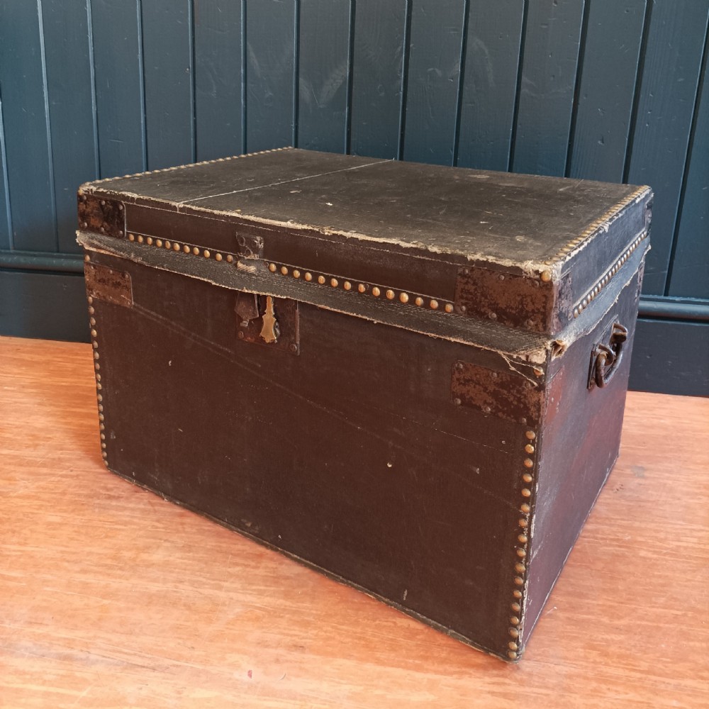 A rectangular dark brown leather campaign trunk featuring brass studding along the lid edges, metal corner brackets, and drop handles.
