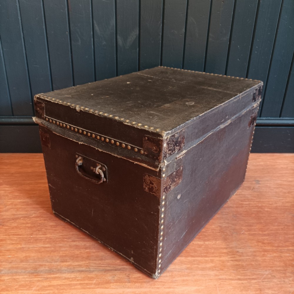 A rectangular dark brown leather campaign trunk featuring brass studding along the lid edges, metal corner brackets, and drop handles. View V