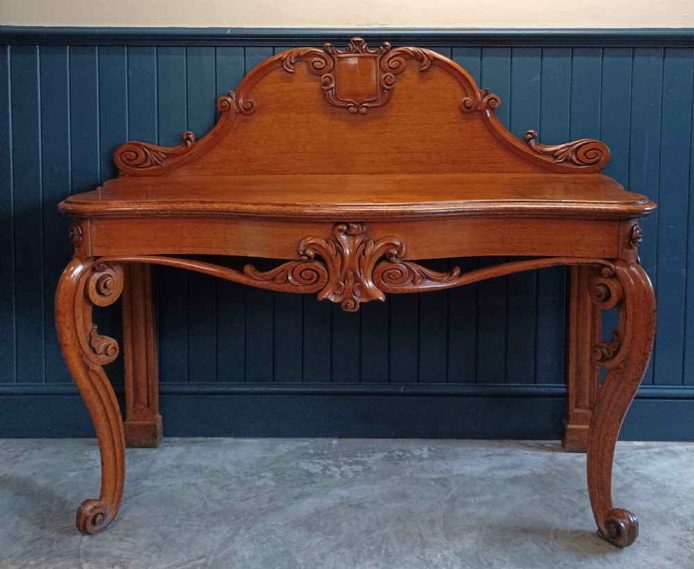 A carved mid-19th-century Victorian oak hall console table featuring scrolled cabriole legs, a central acanthus leaf apron, and a raised gallery back with a carved shield crest.