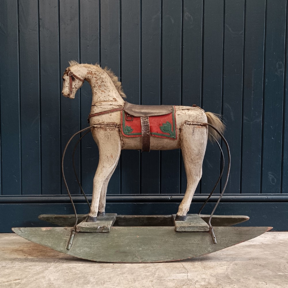 A 19th-century hand-carved wooden rocking horse featuring distressed cream paint, a red saddle cloth, leather tack, and curved green wooden rockers.