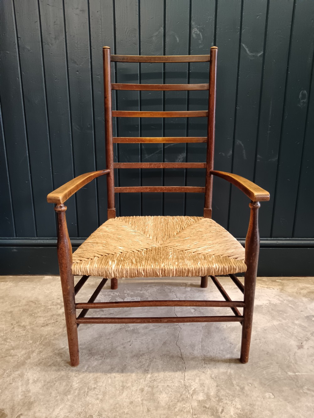 An Arts and Crafts dark wooden ladder back armchair featuring curved armrests and a woven rush seat. View XII
