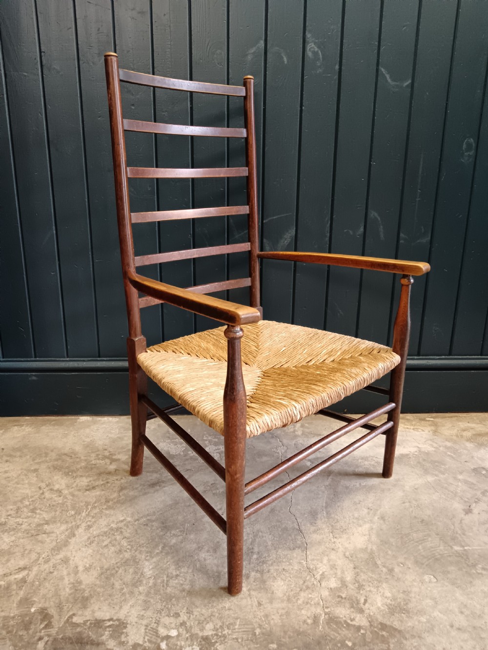 An Arts and Crafts dark wooden ladder back armchair featuring curved armrests and a woven rush seat. View II