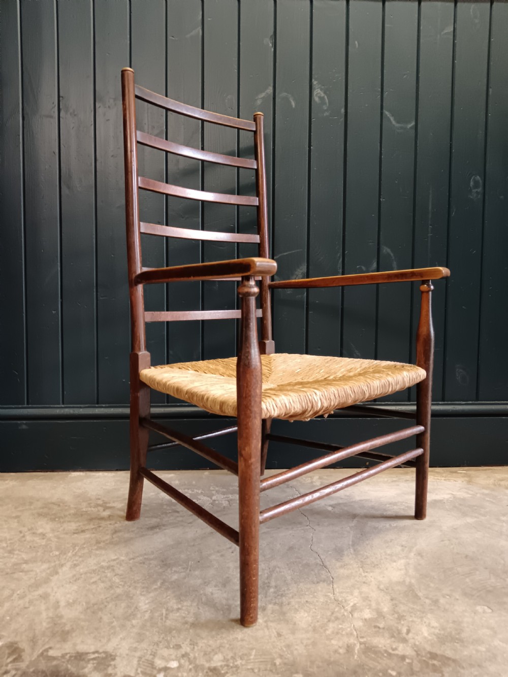 An Arts and Crafts dark wooden ladder back armchair featuring curved armrests and a woven rush seat. View III