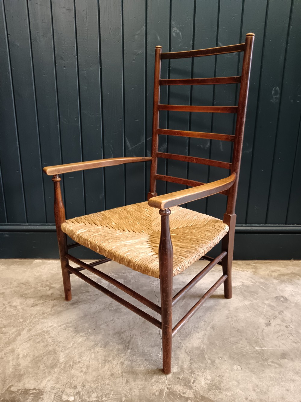 An Arts and Crafts dark wooden ladder back armchair featuring curved armrests and a woven rush seat. View IV