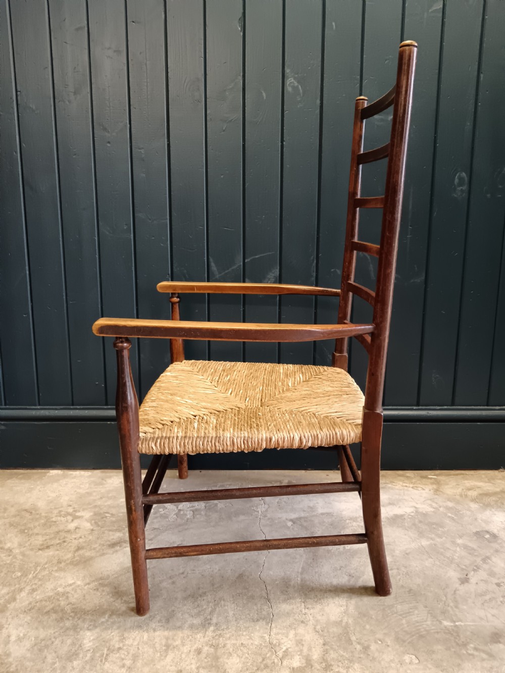 An Arts and Crafts dark wooden ladder back armchair featuring curved armrests and a woven rush seat. View V