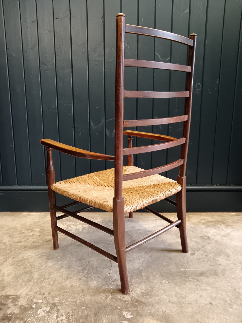 An Arts and Crafts dark wooden ladder back armchair featuring curved armrests and a woven rush seat. View VI