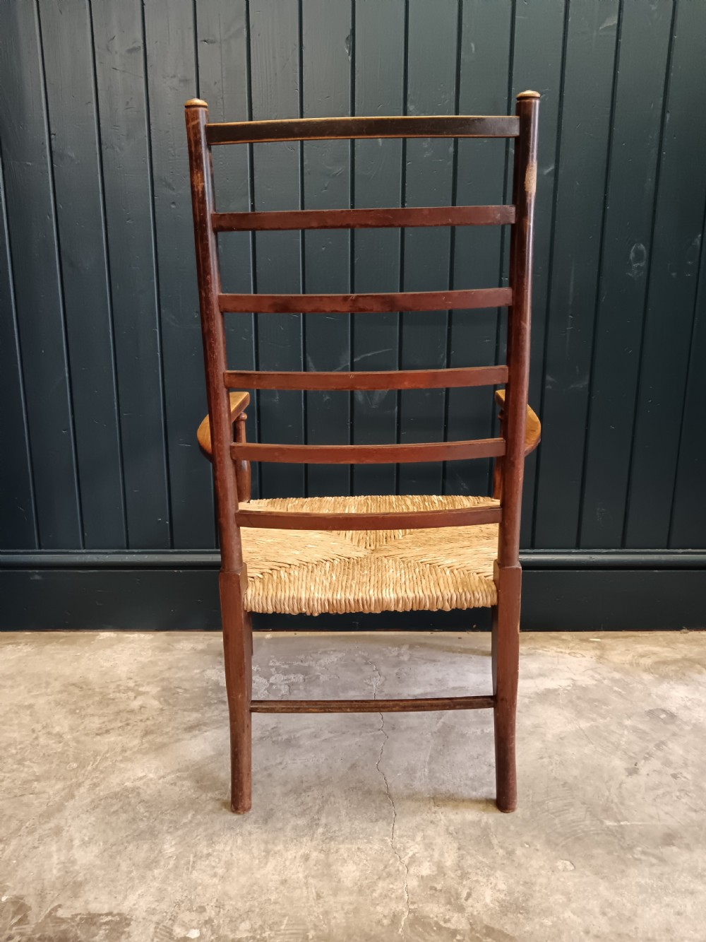 An Arts and Crafts dark wooden ladder back armchair featuring curved armrests and a woven rush seat. View VII