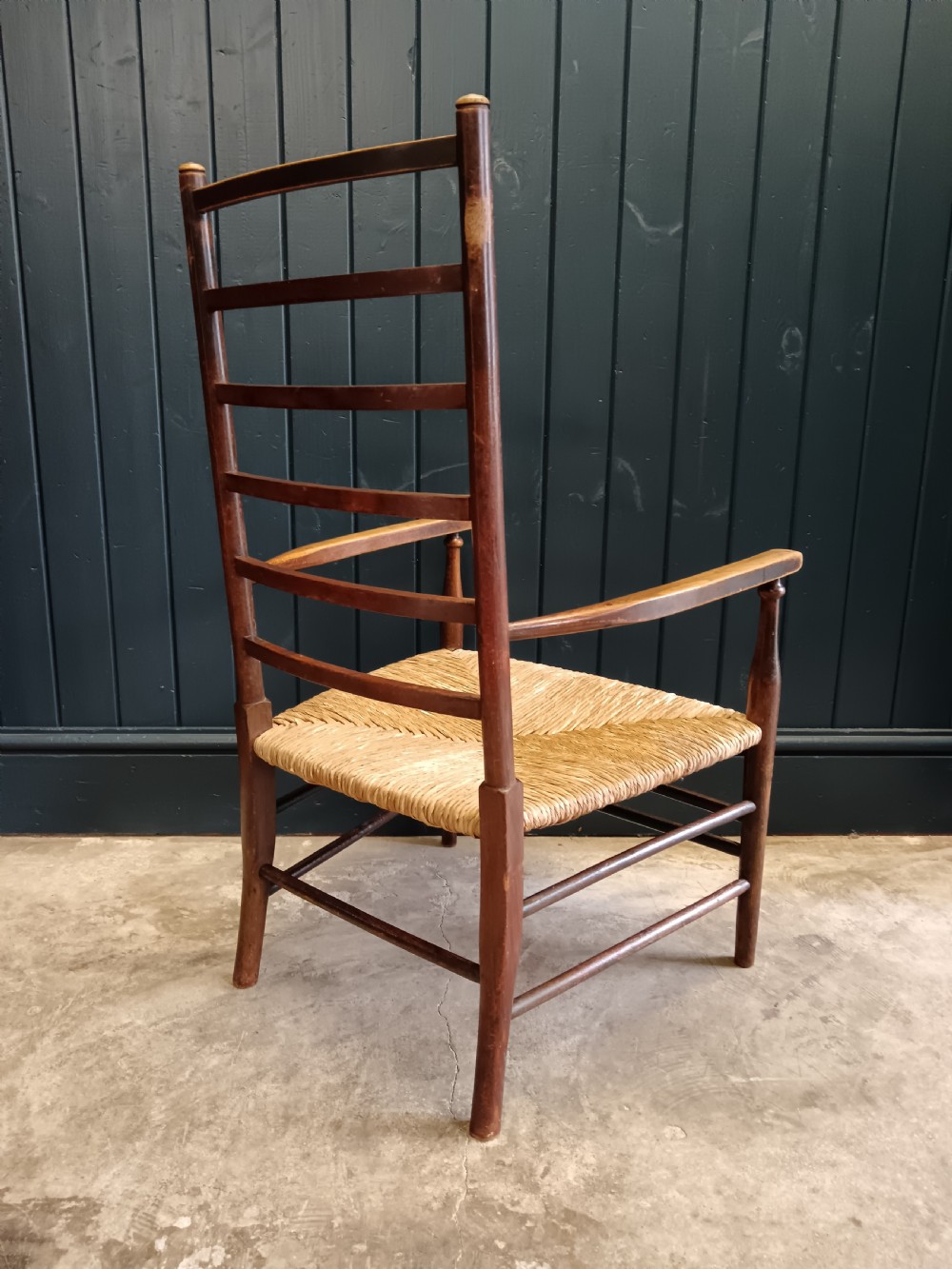 An Arts and Crafts dark wooden ladder back armchair featuring curved armrests and a woven rush seat. View VIII