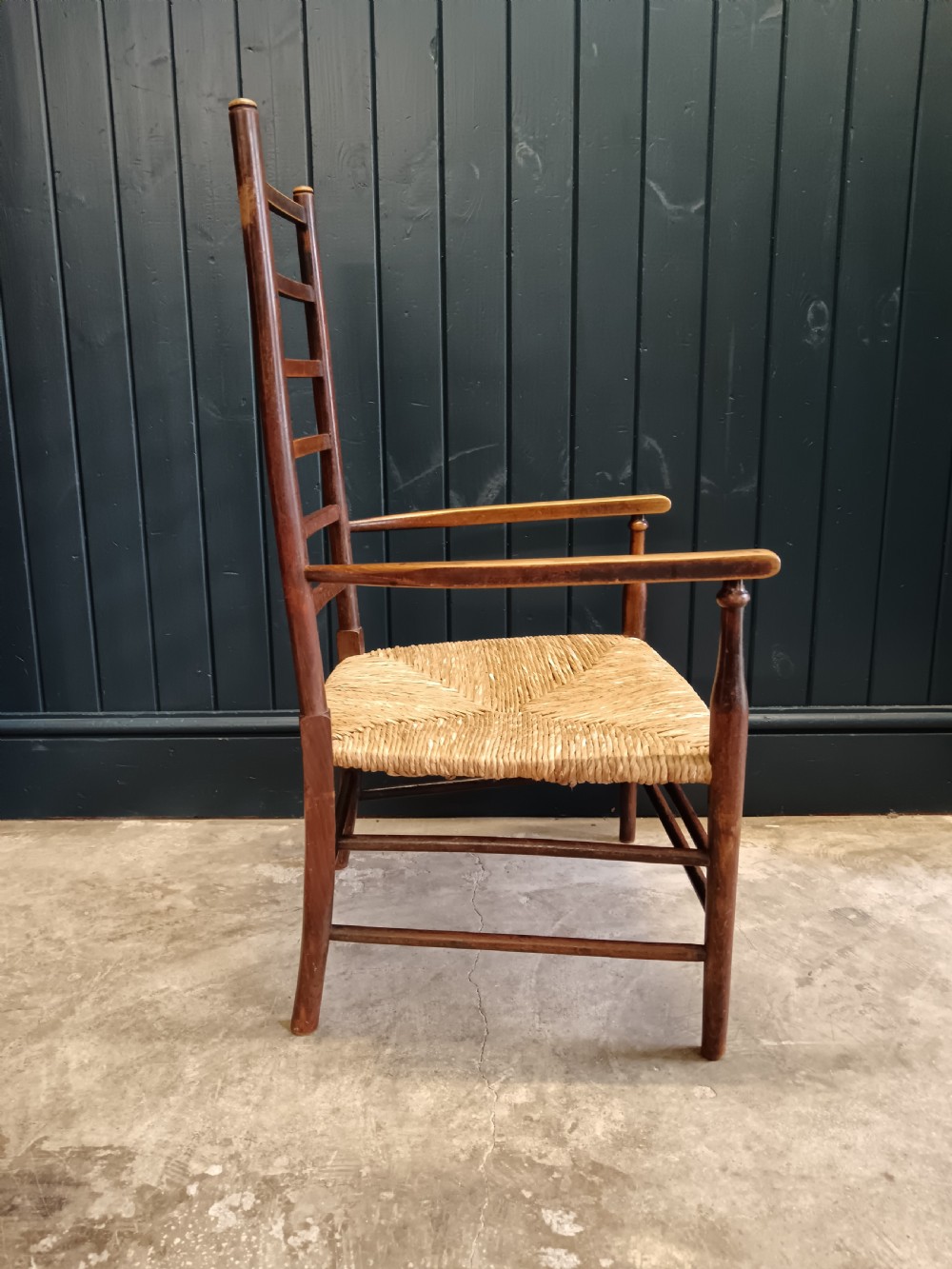 An Arts and Crafts dark wooden ladder back armchair featuring curved armrests and a woven rush seat. View IX