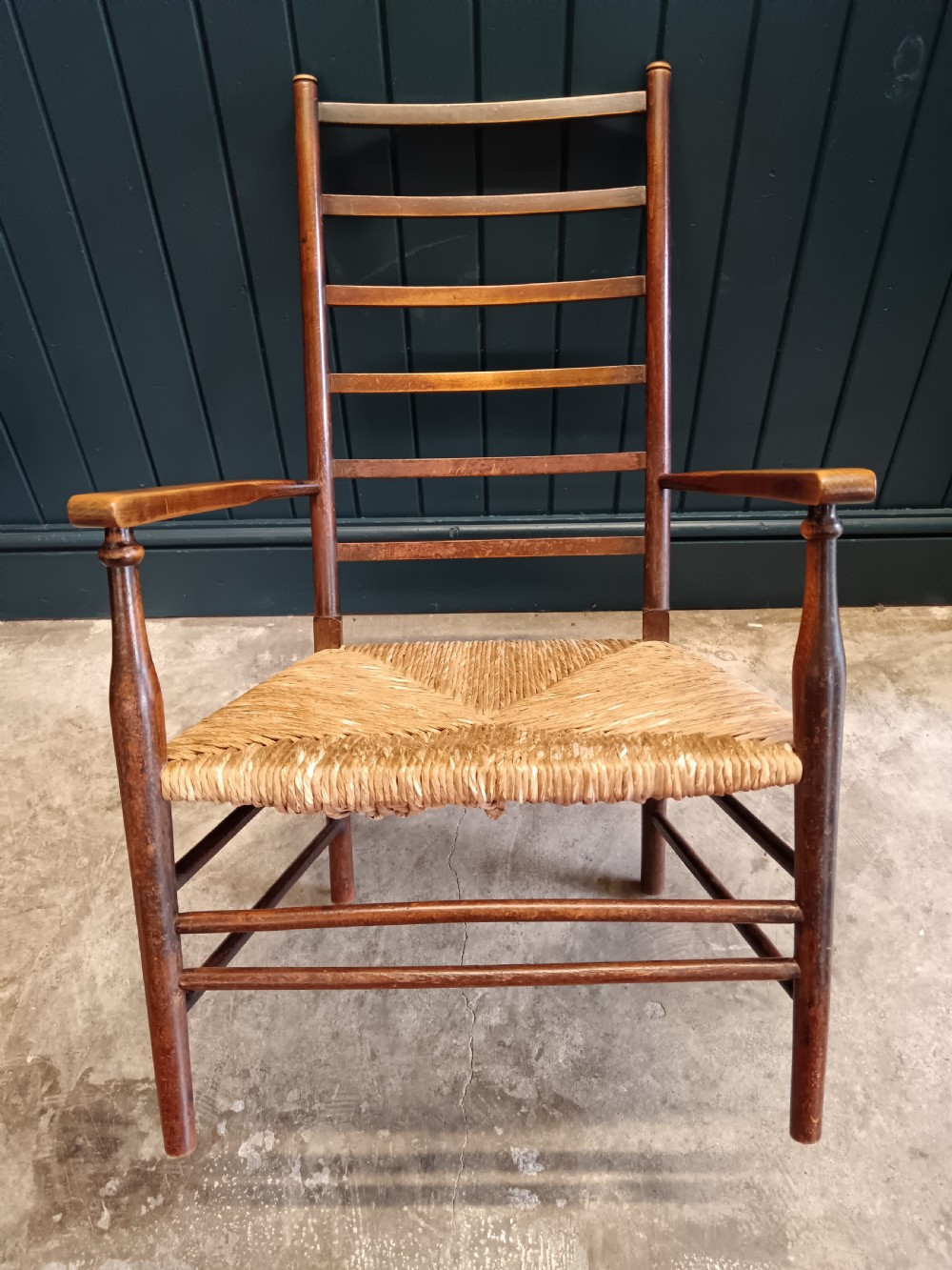 An Arts and Crafts dark wooden ladder back armchair featuring curved armrests and a woven rush seat. View XI