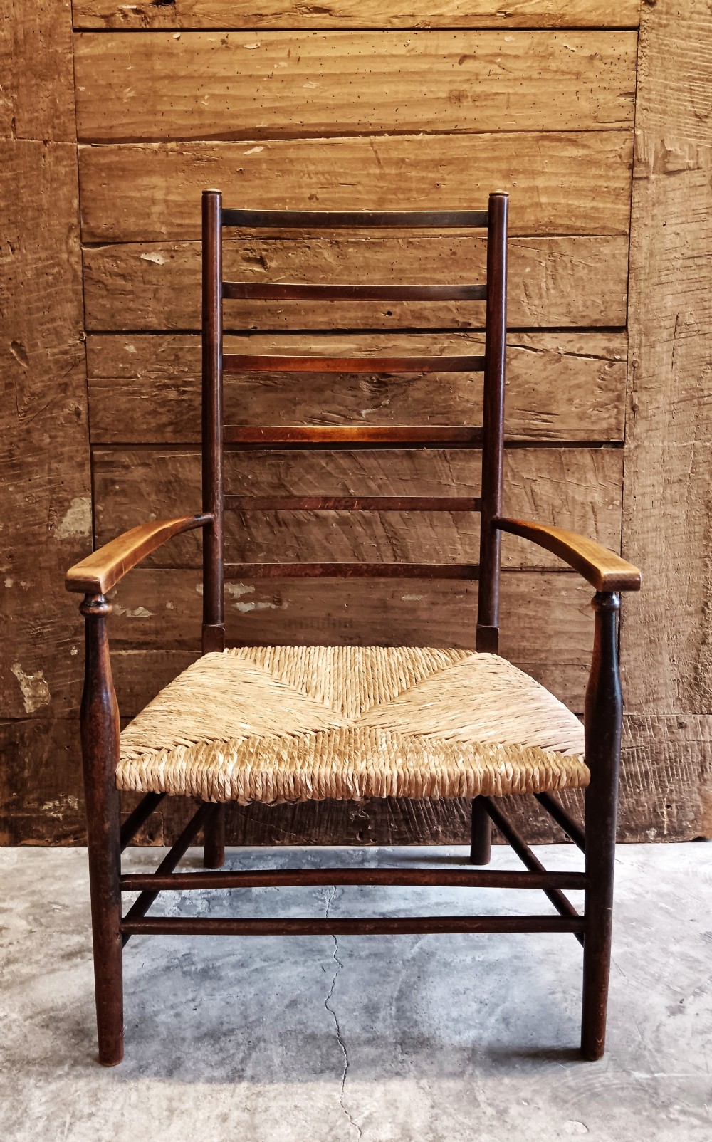 An Arts and Crafts dark wooden ladder back armchair featuring curved armrests and a woven rush seat.