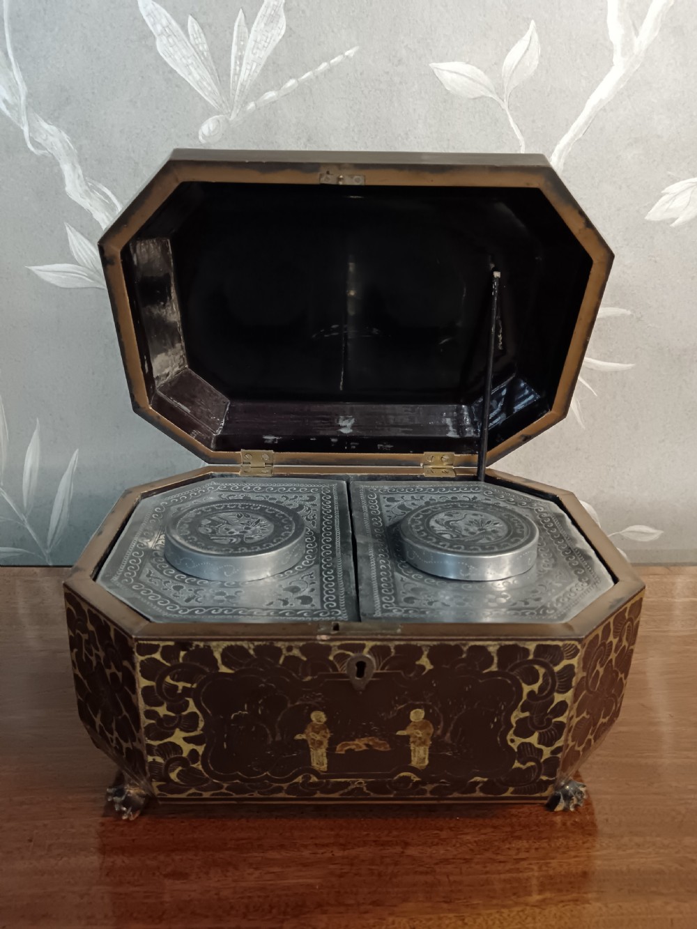 An open octagonal dark lacquer tea box decorated with gilded Chinese motifs containing two engraved pewter metal canisters.