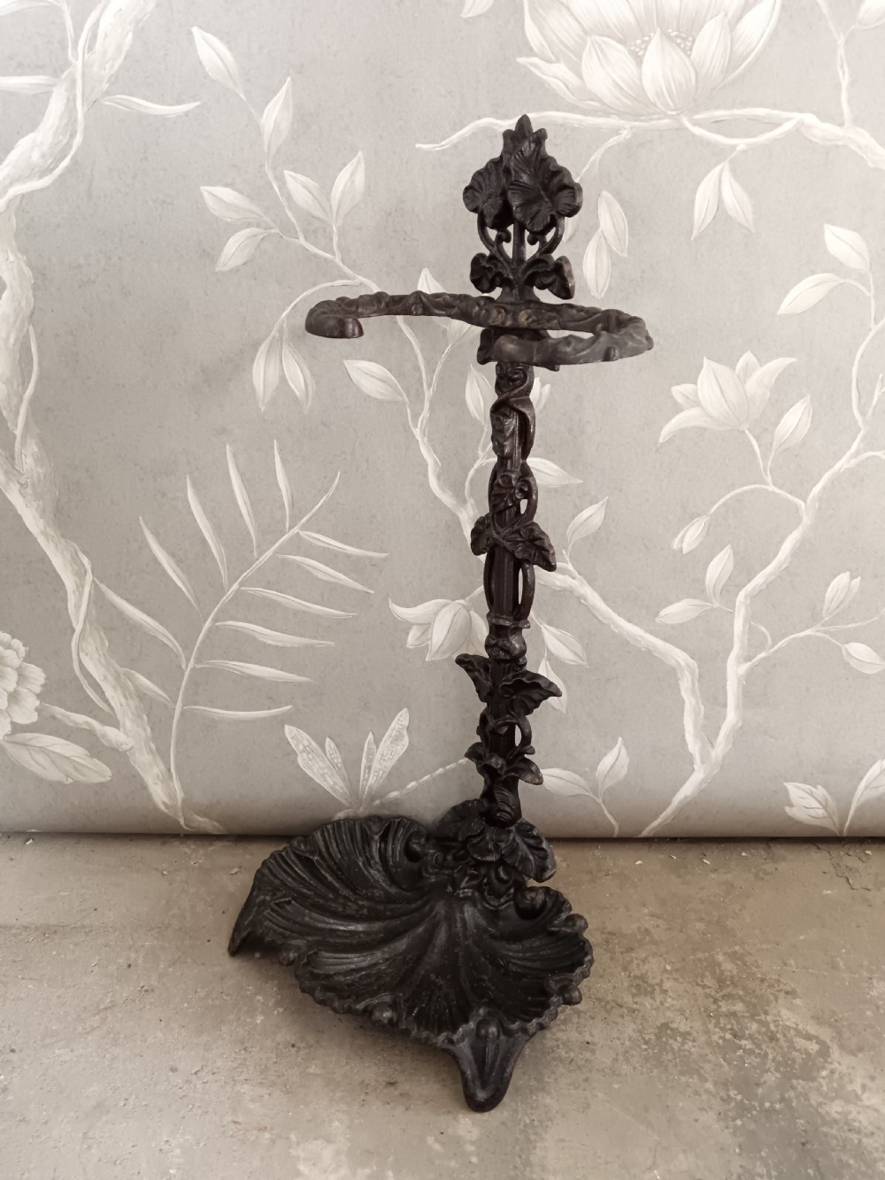 A dark cast iron Art Nouveau umbrella stand featuring stylized floral fretwork, dual upper loops, and a shell-shaped drip tray base. View VI