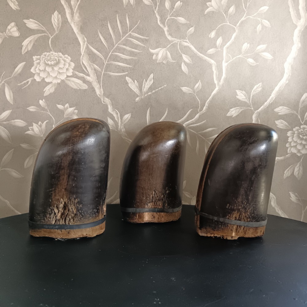 Three tall carved dark wooden millinery hat blocks with sloping rounded tops arranged in a row on a surface. View X