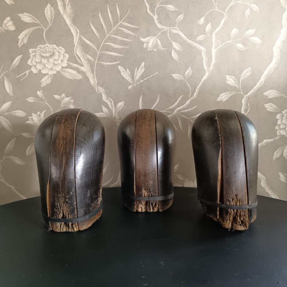 Three tall carved dark wooden millinery hat blocks with sloping rounded tops arranged in a row on a surface. View V