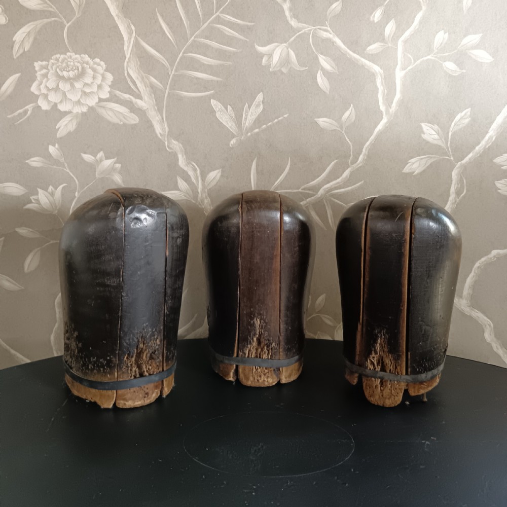 Three tall carved dark wooden millinery hat blocks with sloping rounded tops arranged in a row on a surface. View VII