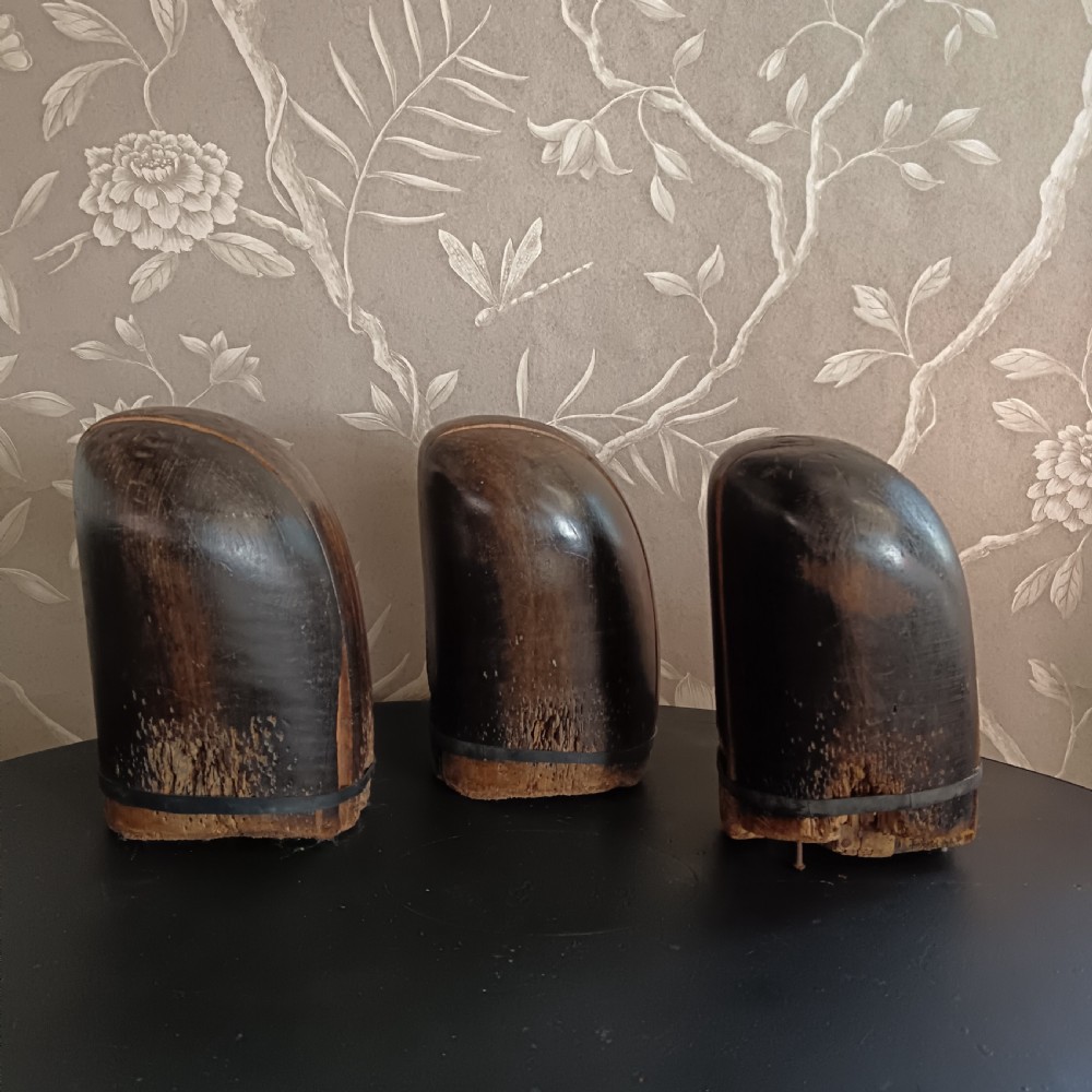 Three tall carved dark wooden millinery hat blocks with sloping rounded tops arranged in a row on a surface.