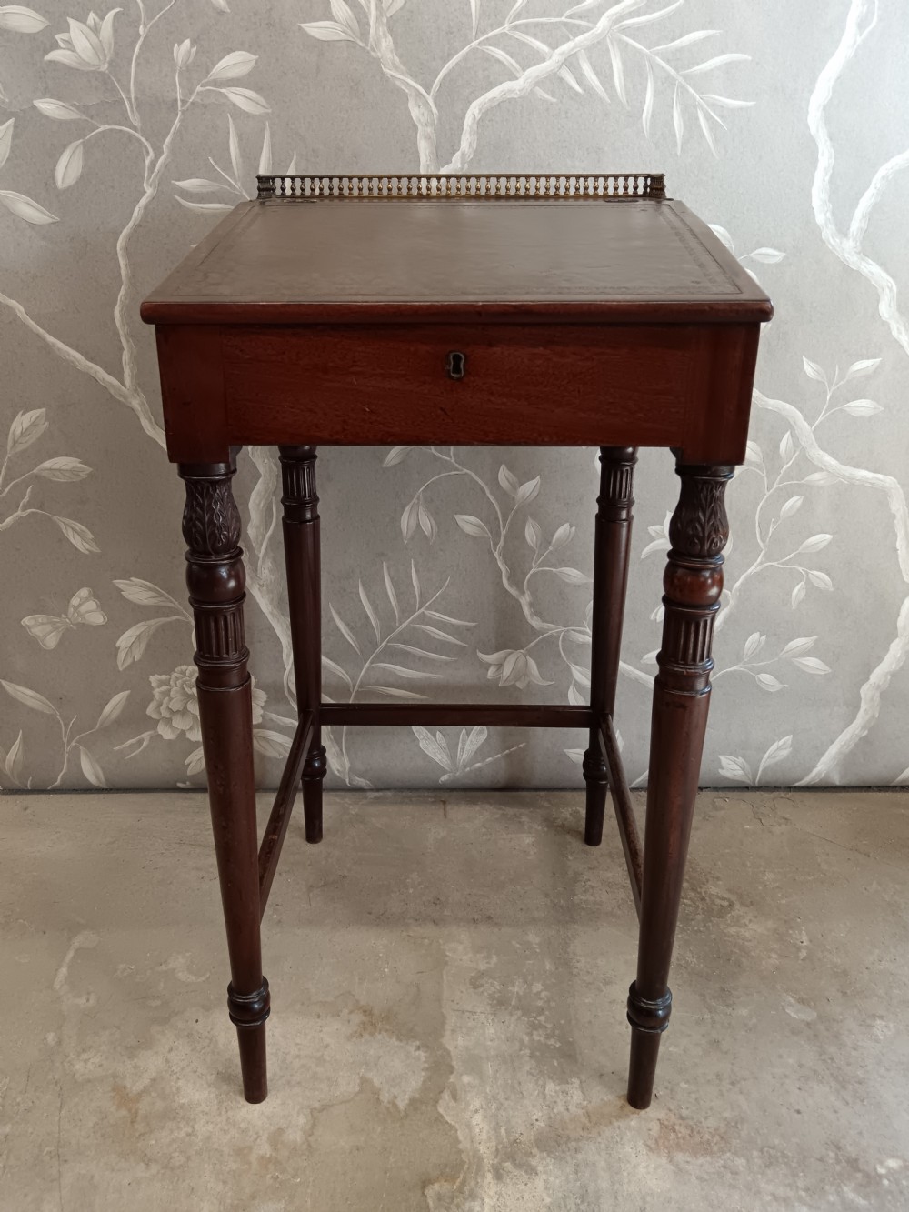 A dark mahogany Regency ladys writing desk featuring turned reeded legs, a single drawer with a keyhole escutcheon, a leather inset top, and a brass gallery along the rear edge.