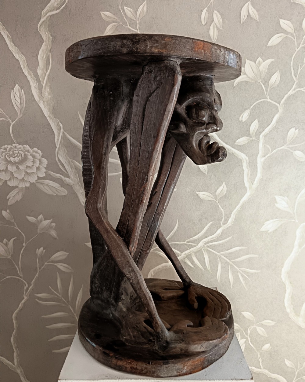 A dark carved timber side table featuring a circular top supported by a stylised crouching figure above a rounded base. View VII