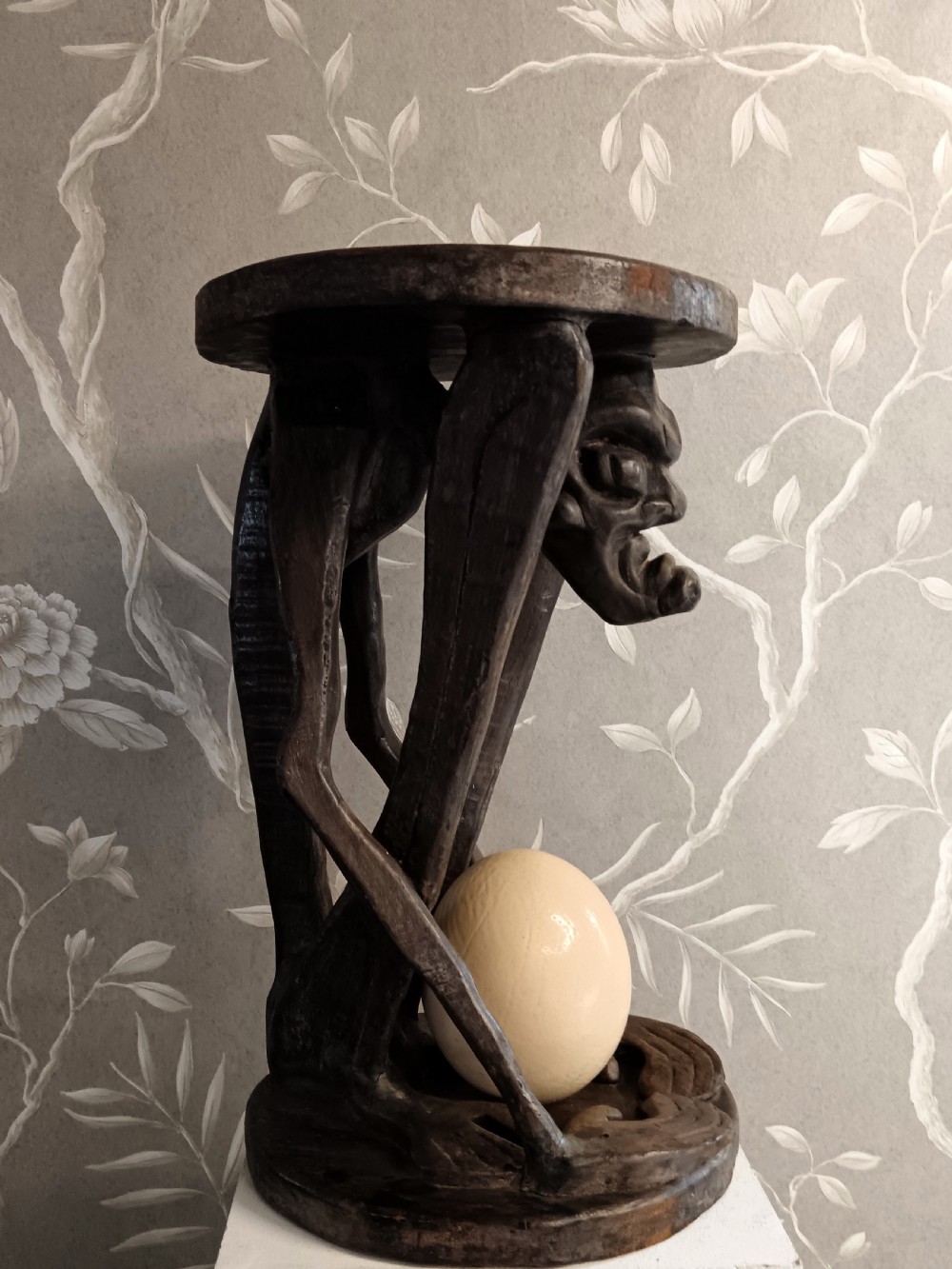 A dark carved timber side table featuring a circular top supported by a stylised crouching figure above a rounded base.