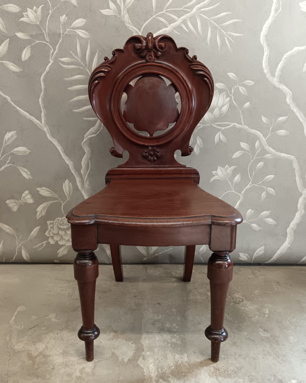 A Victorian mahogany hall chair featuring a carved scrollwork backrest with a central shield motif, a solid timber seat, and turned front legs.