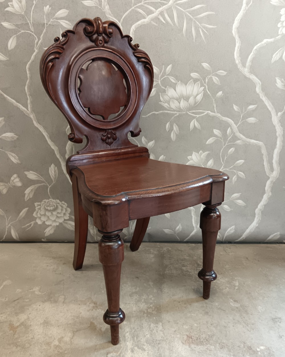 A Victorian mahogany hall chair featuring a carved scrollwork backrest with a central shield motif, a solid timber seat, and turned front legs. View II
