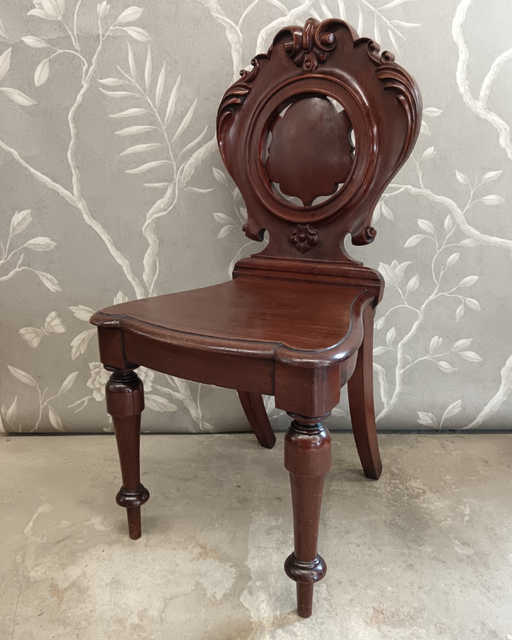 A Victorian mahogany hall chair featuring a carved scrollwork backrest with a central shield motif, a solid timber seat, and turned front legs. View III