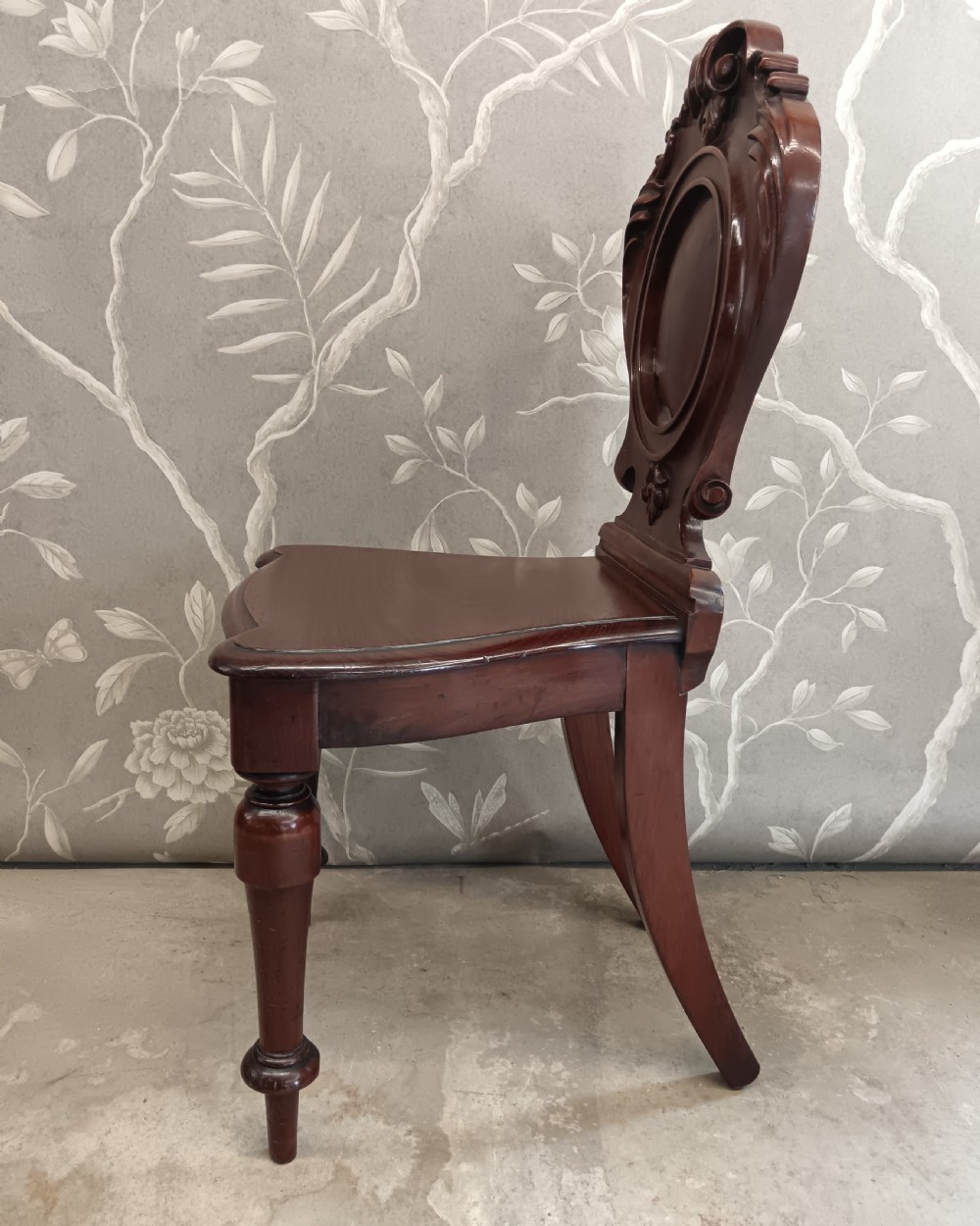 A Victorian mahogany hall chair featuring a carved scrollwork backrest with a central shield motif, a solid timber seat, and turned front legs. View IV