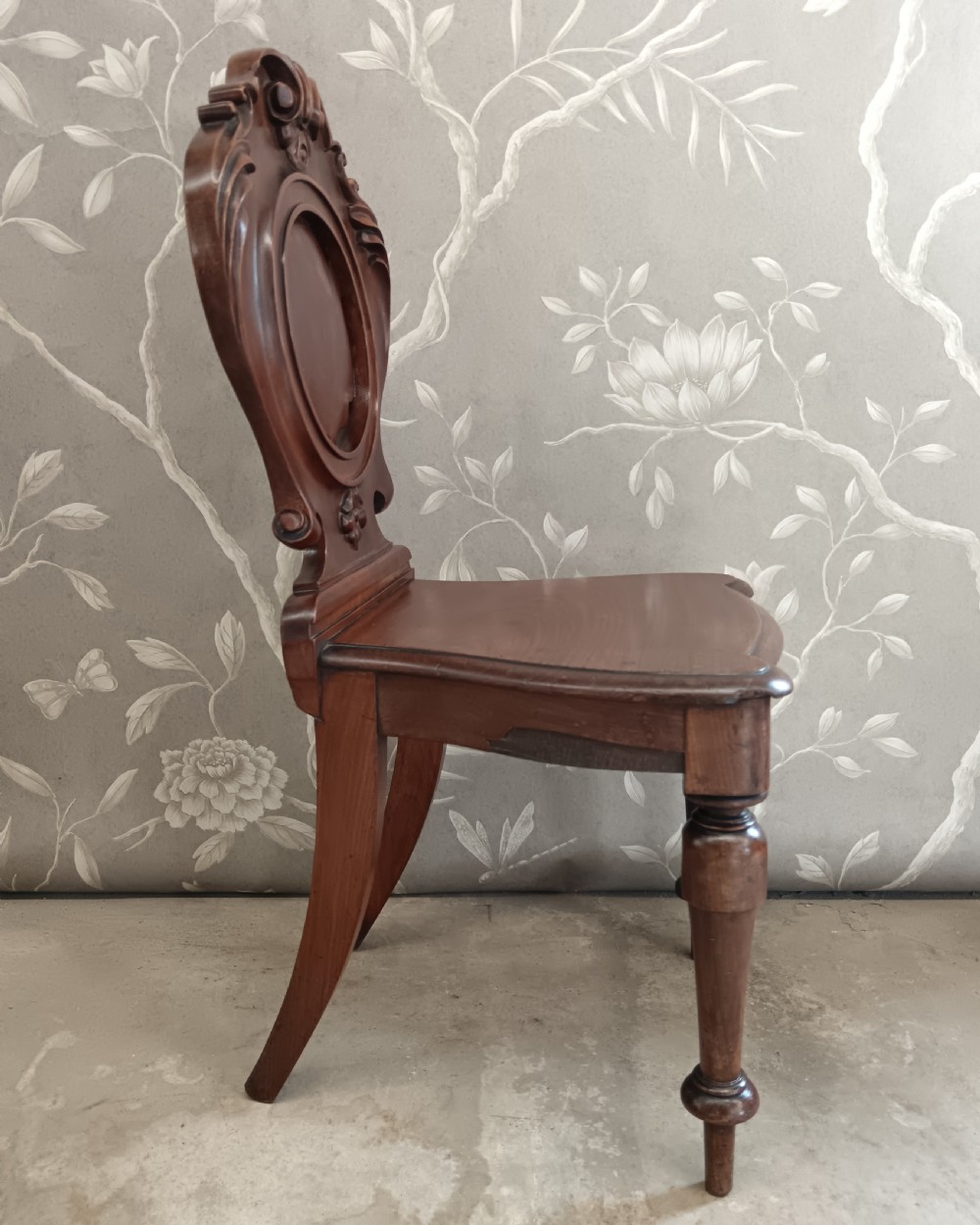 A Victorian mahogany hall chair featuring a carved scrollwork backrest with a central shield motif, a solid timber seat, and turned front legs. View V