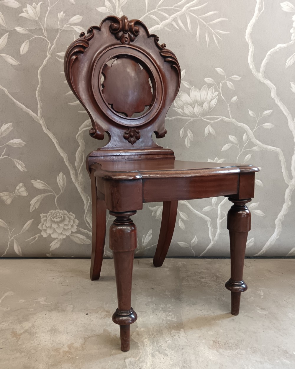 A Victorian mahogany hall chair featuring a carved scrollwork backrest with a central shield motif, a solid timber seat, and turned front legs. View VII